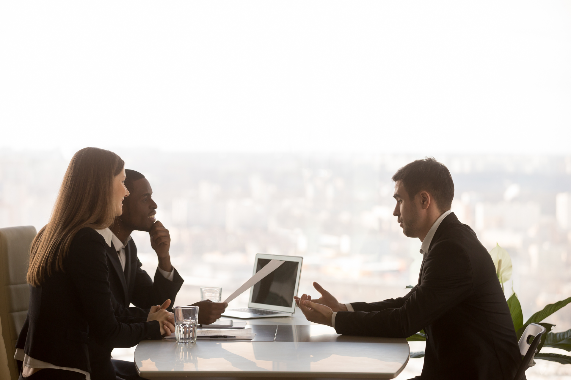 Friendly attentive hr managers interviewing vacancy applicant, multi-ethnic partners discussing new project idea sitting at office desk with cityscape outside